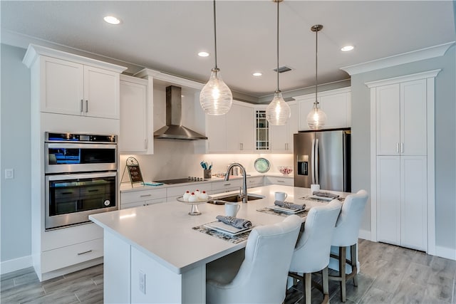 kitchen with sink, hanging light fixtures, stainless steel appliances, wall chimney range hood, and an island with sink