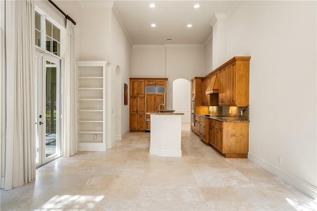 kitchen featuring arched walkways, brown cabinetry, a high ceiling, and a kitchen island with sink