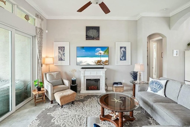 living area with arched walkways, ceiling fan, a warm lit fireplace, tile patterned flooring, and ornamental molding