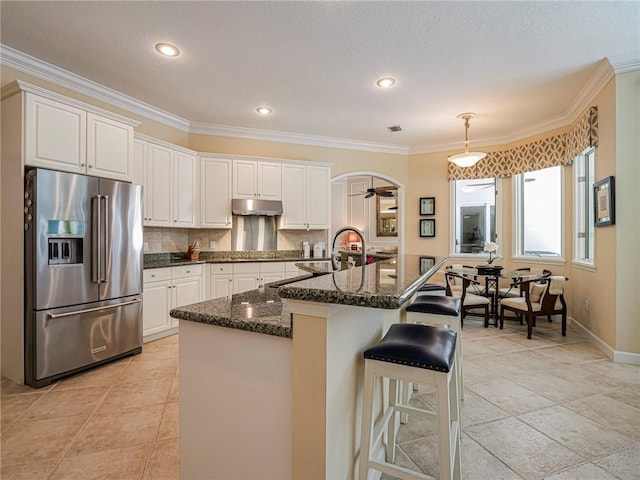 kitchen featuring high quality fridge, tasteful backsplash, white cabinets, dark stone counters, and a center island with sink