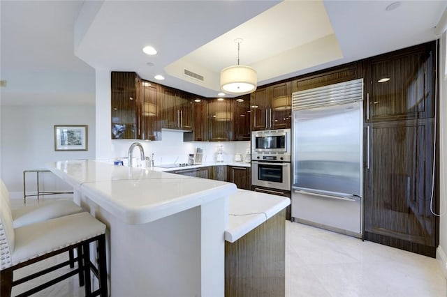 kitchen featuring built in appliances, a peninsula, a raised ceiling, light countertops, and a breakfast bar