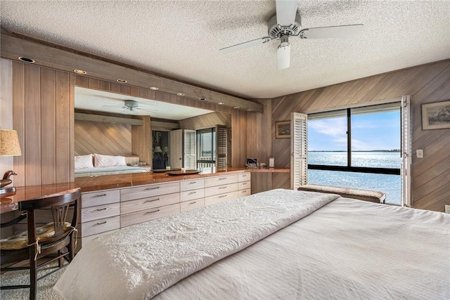 bedroom with a textured ceiling, ceiling fan, a water view, and wood walls