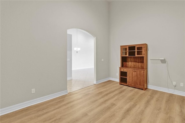 empty room featuring a towering ceiling, arched walkways, baseboards, an inviting chandelier, and light wood-style flooring