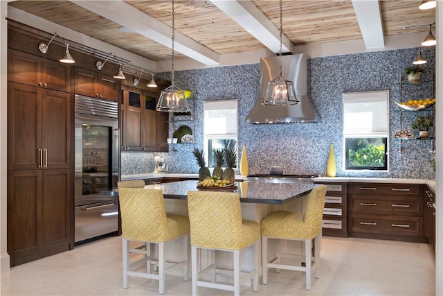 kitchen with pendant lighting, wall chimney range hood, stainless steel built in refrigerator, tasteful backsplash, and beamed ceiling