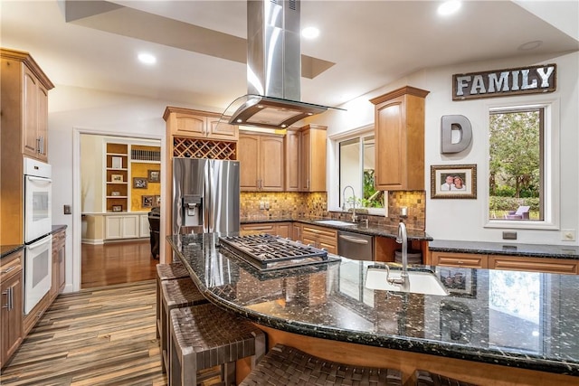 kitchen with stainless steel appliances, dark stone countertops, a sink, and island range hood