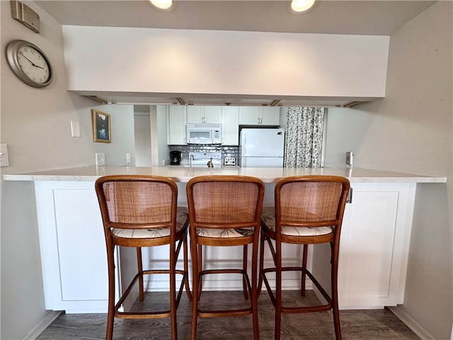 kitchen featuring dark wood-type flooring, a kitchen breakfast bar, kitchen peninsula, white appliances, and white cabinets