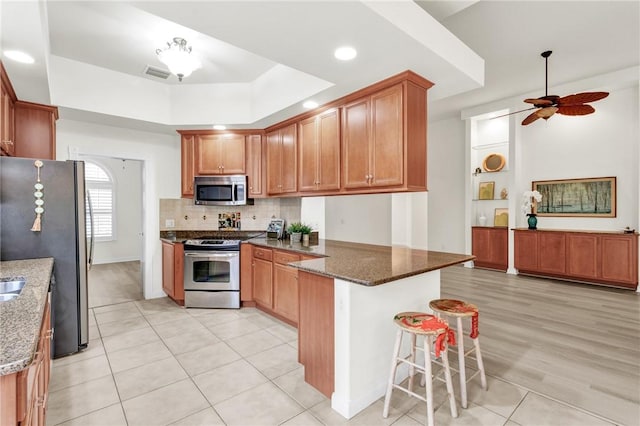 kitchen featuring appliances with stainless steel finishes, dark stone countertops, kitchen peninsula, a breakfast bar, and a tray ceiling