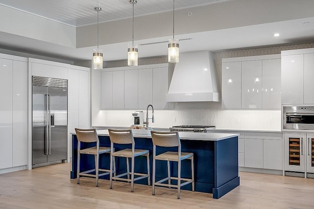 kitchen with a center island with sink, hanging light fixtures, appliances with stainless steel finishes, custom range hood, and white cabinets