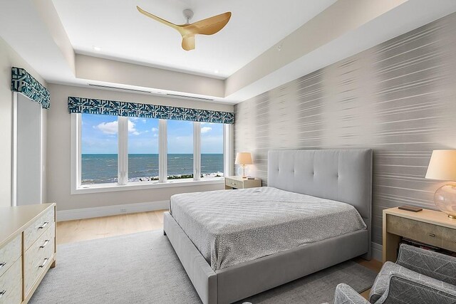 bedroom with a water view, light hardwood / wood-style flooring, ceiling fan, and a tray ceiling