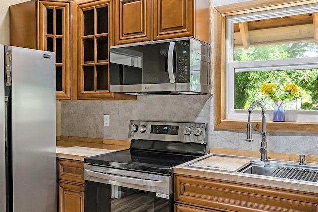 kitchen featuring light countertops, brown cabinetry, stainless steel appliances, decorative backsplash, and glass insert cabinets