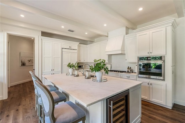 kitchen with beam ceiling, beverage cooler, an island with sink, white cabinets, and appliances with stainless steel finishes