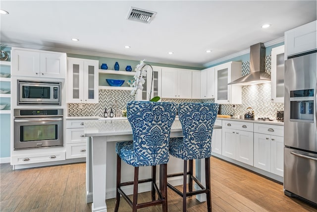 kitchen with white cabinets, appliances with stainless steel finishes, wall chimney exhaust hood, and light hardwood / wood-style floors