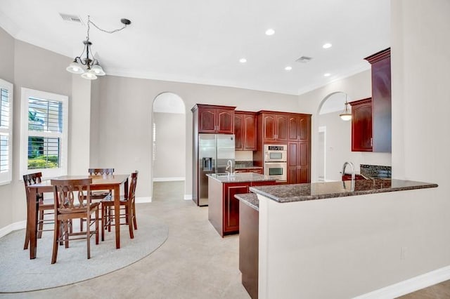 kitchen featuring visible vents, an island with sink, arched walkways, stainless steel appliances, and dark brown cabinets