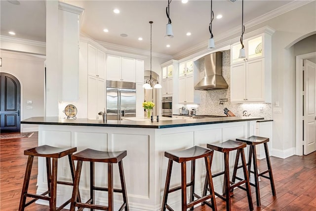 kitchen with built in refrigerator, white cabinetry, a breakfast bar area, and wall chimney range hood