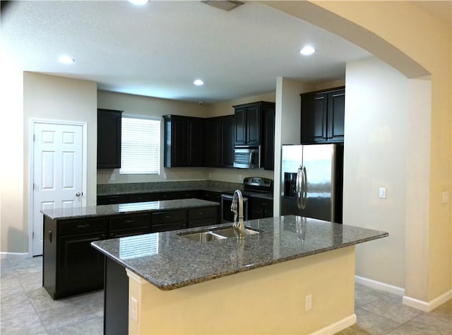 kitchen with arched walkways, a kitchen island with sink, stainless steel appliances, a sink, and dark stone counters