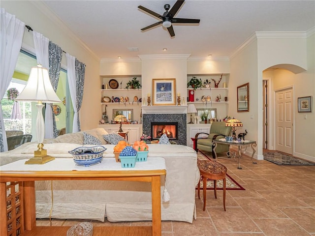 living area featuring a warm lit fireplace, ornamental molding, built in shelves, a ceiling fan, and arched walkways