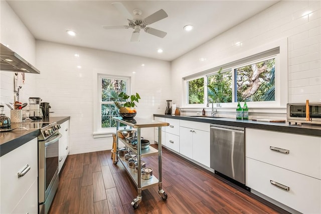 kitchen featuring a sink, dark countertops, appliances with stainless steel finishes, wall chimney exhaust hood, and white cabinets