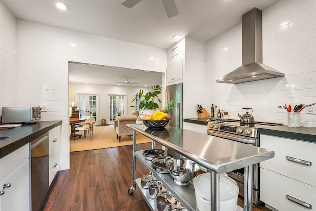 kitchen featuring dark countertops, wall chimney range hood, appliances with stainless steel finishes, white cabinets, and a ceiling fan