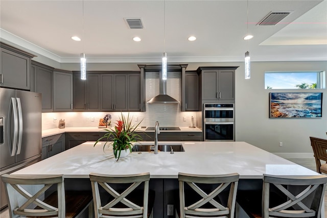 kitchen with a kitchen island with sink, hanging light fixtures, stainless steel appliances, and wall chimney range hood