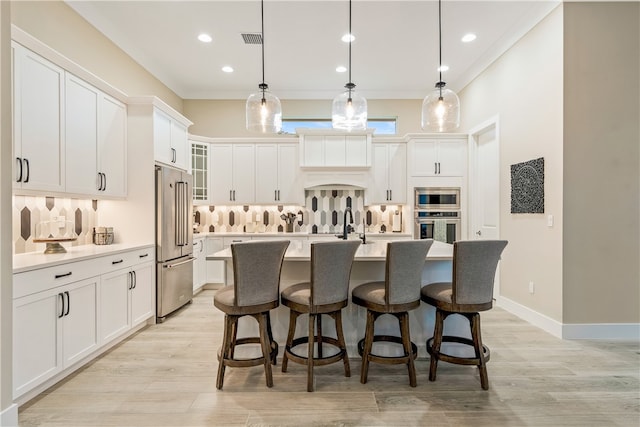 kitchen with stainless steel appliances, a kitchen island with sink, decorative light fixtures, white cabinets, and light hardwood / wood-style floors