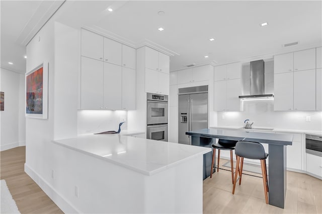 kitchen featuring stainless steel appliances, light hardwood / wood-style floors, wall chimney exhaust hood, and a breakfast bar