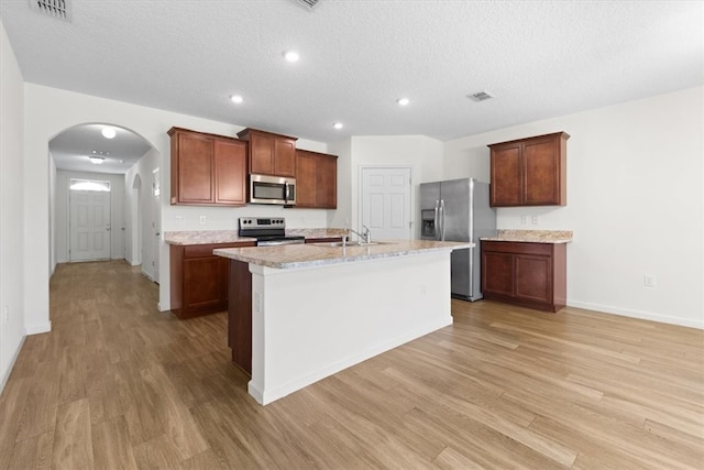 kitchen with a textured ceiling, light wood-type flooring, stainless steel appliances, and a kitchen island with sink