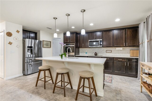 kitchen featuring backsplash, appliances with stainless steel finishes, decorative light fixtures, dark brown cabinetry, and a kitchen island with sink