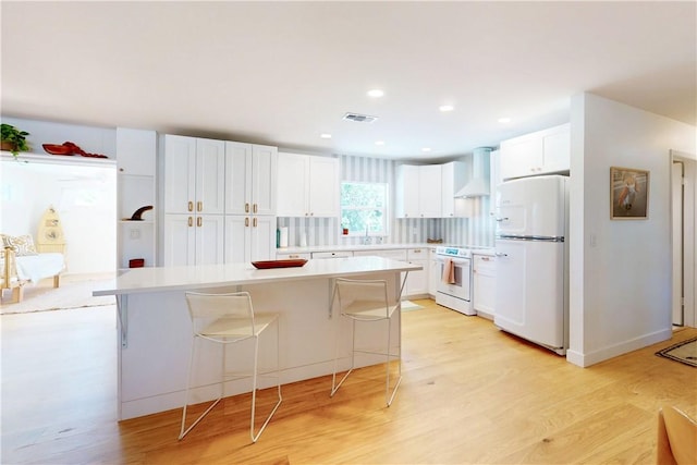 kitchen with a kitchen breakfast bar, white appliances, white cabinets, wall chimney range hood, and a kitchen island