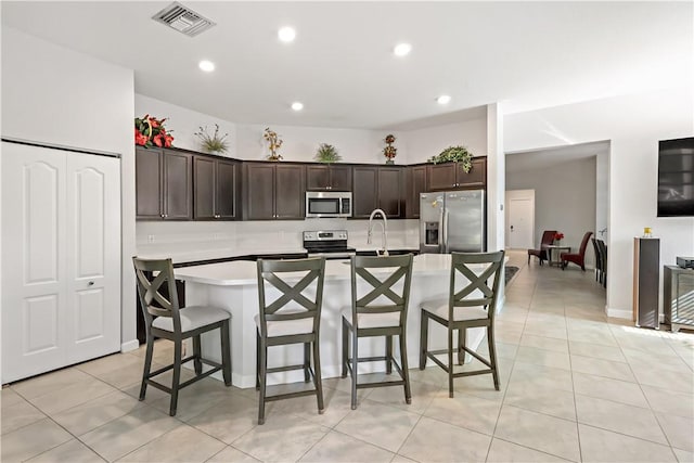 kitchen featuring appliances with stainless steel finishes, dark brown cabinets, light tile patterned floors, a breakfast bar area, and an island with sink