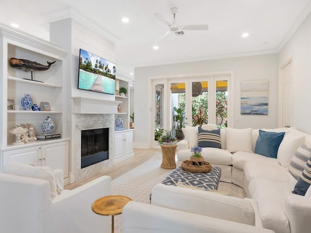 living room featuring built in features, ceiling fan, a high end fireplace, crown molding, and light wood-type flooring