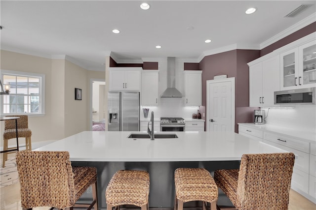 kitchen featuring a center island with sink, sink, wall chimney range hood, white cabinetry, and appliances with stainless steel finishes