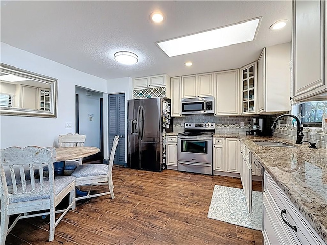 kitchen with a sink, light stone counters, dark wood finished floors, stainless steel appliances, and glass insert cabinets