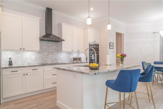 kitchen with white cabinetry, wall chimney range hood, crown molding, and a kitchen island with sink