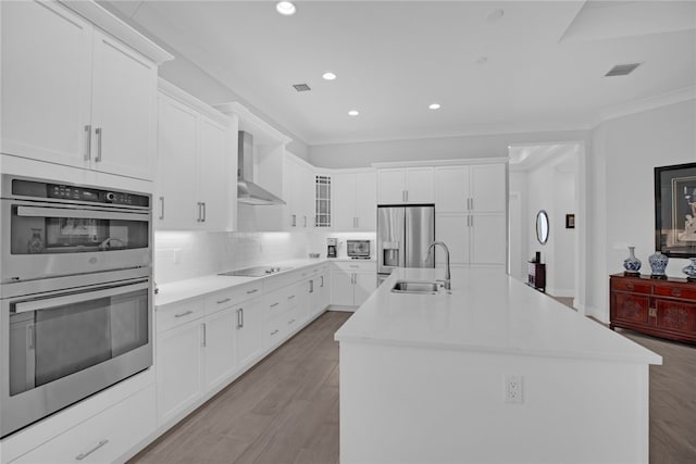 kitchen with stainless steel appliances, white cabinetry, wall chimney exhaust hood, an island with sink, and light hardwood / wood-style flooring