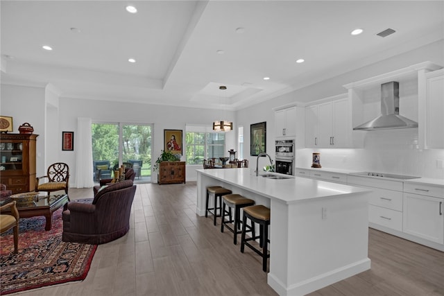 kitchen with white cabinetry, wall chimney range hood, and an island with sink