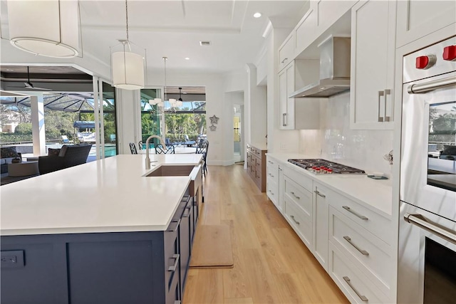 kitchen with white cabinetry, appliances with stainless steel finishes, an island with sink, pendant lighting, and wall chimney range hood