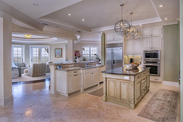 kitchen featuring built in appliances, hanging light fixtures, a kitchen island with sink, a tray ceiling, and dark stone countertops