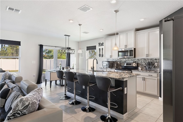 kitchen with white cabinetry, light stone countertops, hanging light fixtures, an island with sink, and appliances with stainless steel finishes