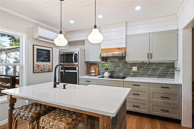 kitchen with gray cabinets, appliances with stainless steel finishes, a wall mounted air conditioner, an island with sink, and hanging light fixtures