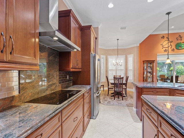 kitchen featuring black electric cooktop, wall chimney range hood, a chandelier, stainless steel refrigerator, and hanging light fixtures