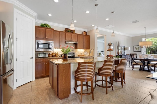 kitchen with light stone countertops, appliances with stainless steel finishes, a kitchen island with sink, crown molding, and decorative light fixtures