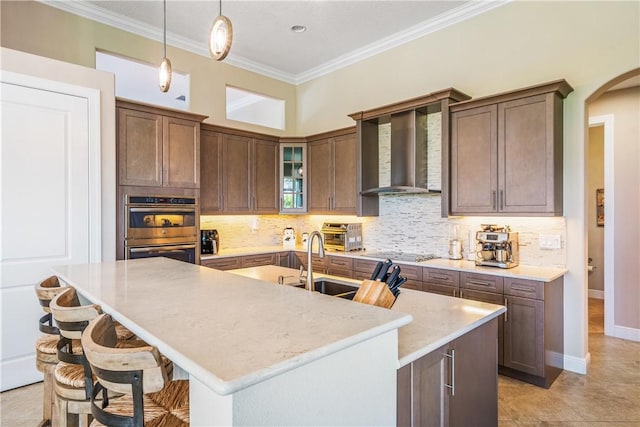 kitchen featuring double oven, a kitchen island with sink, light stone counters, black cooktop, and wall chimney exhaust hood