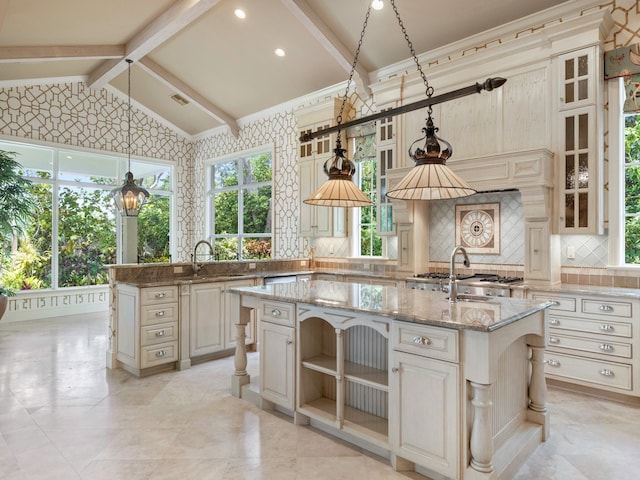 kitchen featuring tasteful backsplash, light stone counters, vaulted ceiling with beams, a center island with sink, and decorative light fixtures