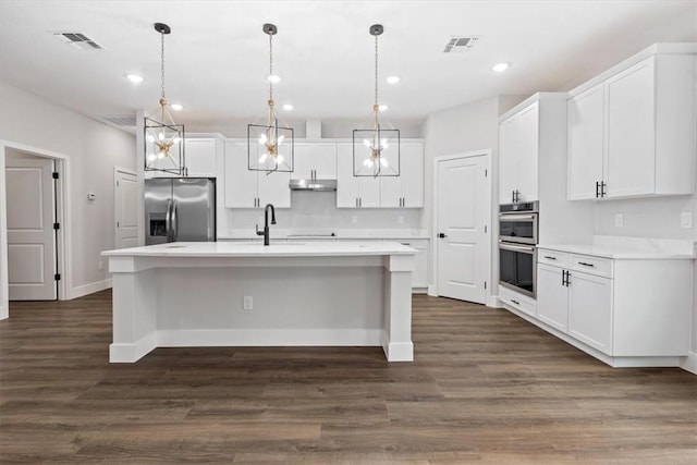 kitchen featuring appliances with stainless steel finishes, a center island with sink, decorative light fixtures, dark hardwood / wood-style floors, and white cabinetry