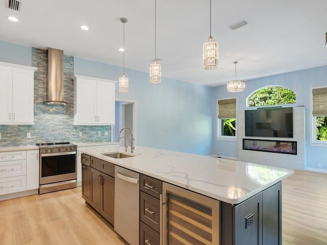 kitchen with wall chimney range hood, appliances with stainless steel finishes, wine cooler, white cabinets, and a center island with sink