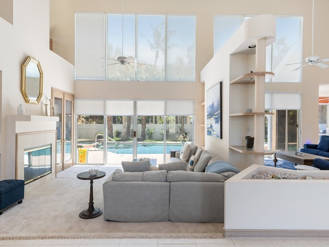 living room featuring a towering ceiling, built in shelves, ceiling fan, a tile fireplace, and light tile patterned floors