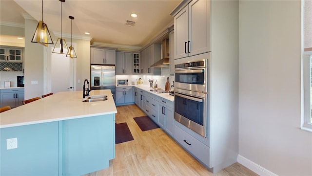 kitchen featuring a center island with sink, stainless steel appliances, wall chimney range hood, decorative light fixtures, and sink