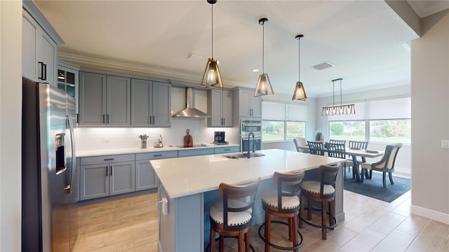 kitchen featuring wall chimney range hood, appliances with stainless steel finishes, hanging light fixtures, a breakfast bar area, and a kitchen island with sink