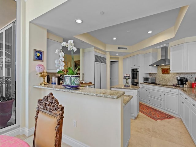 kitchen with tasteful backsplash, visible vents, wall chimney range hood, a tray ceiling, and light stone countertops