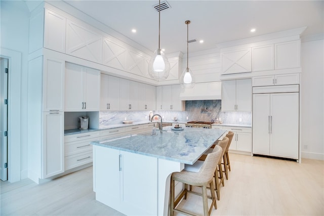 kitchen with light stone countertops, a kitchen island with sink, paneled built in fridge, white cabinets, and pendant lighting